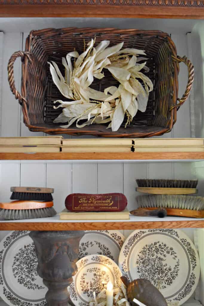 Cornhusk wreath on wicker basket with old books, brushes and plates in a fall display.