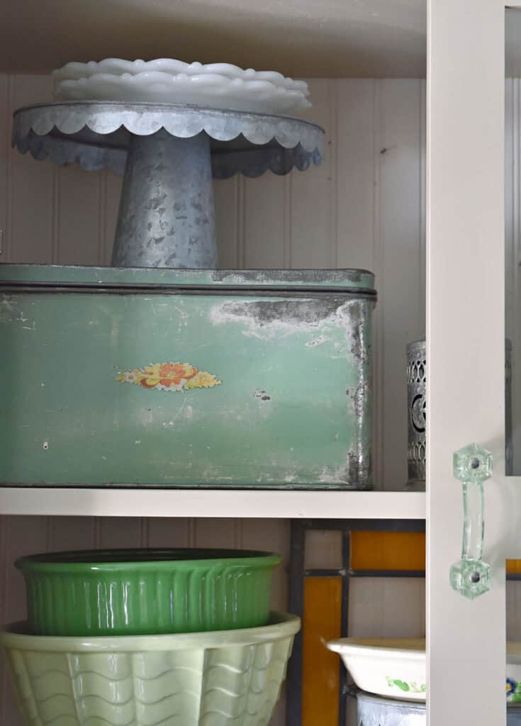 Green metal box, green mixing bowls and silver cake plate in kitchen cupboards.
