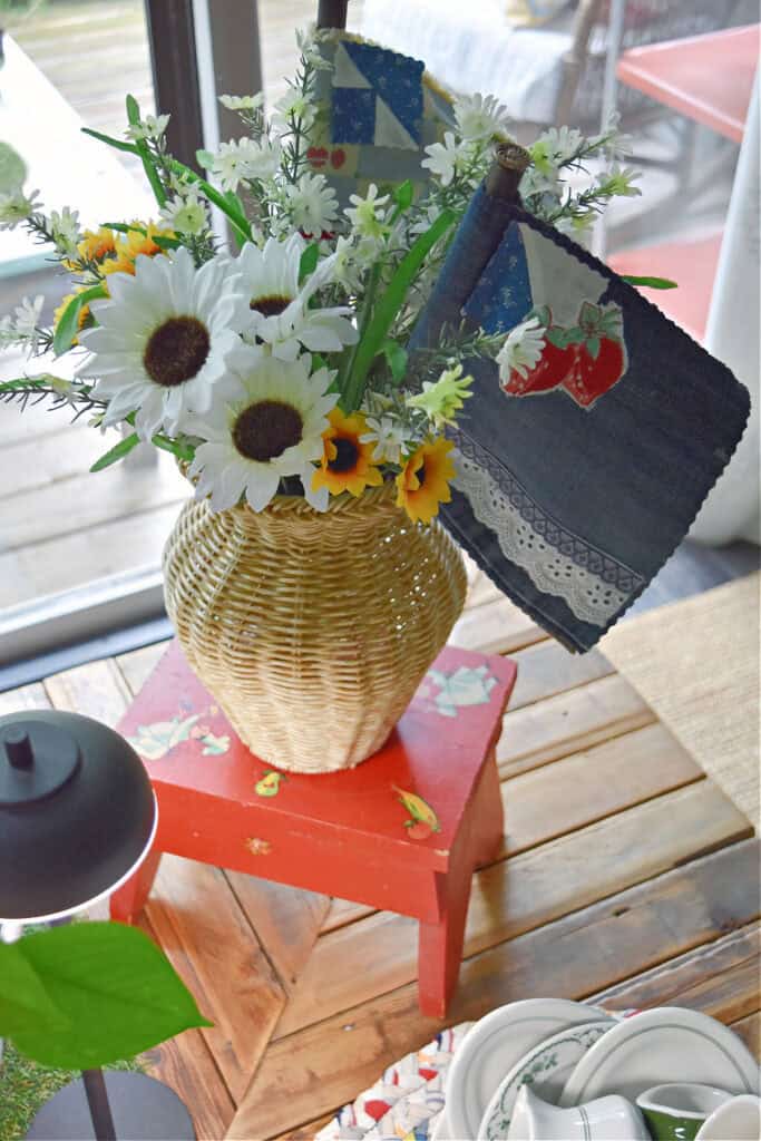 Wicker vase with sunflowers, denim flag on red stool.