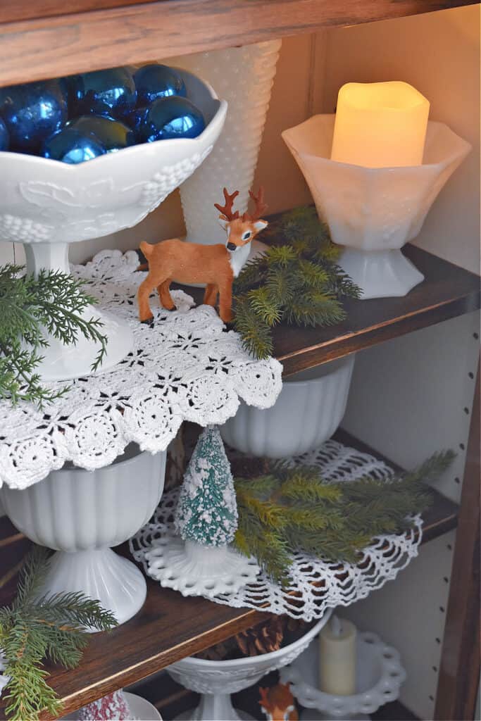 Vintage milk glass and crochet lace and pinecones in bookshelf for Christmas.