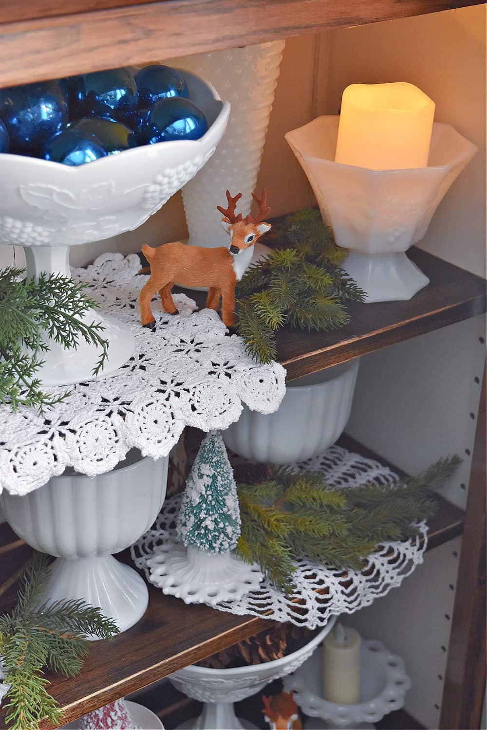 Vintage milk glass and crochet lace and pinecones in bookshelf for Christmas.