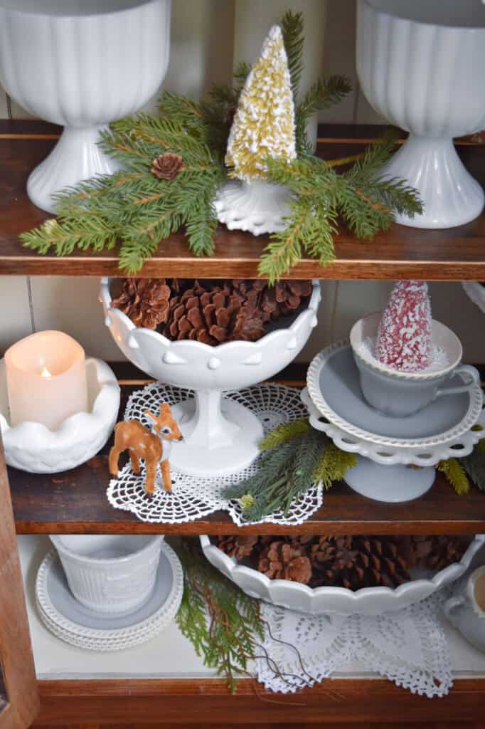 Vintage milk glass and crochet lace and pinecones in bookshelf for Christmas.